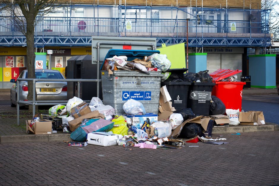 A large collection of mixed household waste and rubbish, including crumpled cardboard boxes, plastic bags filled with waste, paper packaging, and various discarded items, is piled unevenly on a paved