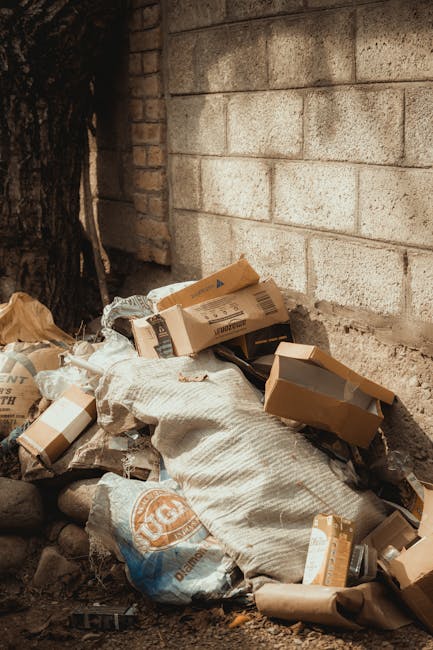 A large collection of mixed household waste and rubbish, including crumpled cardboard boxes, plastic bags filled with waste, paper packaging, and various discarded items, is piled unevenly on a paved